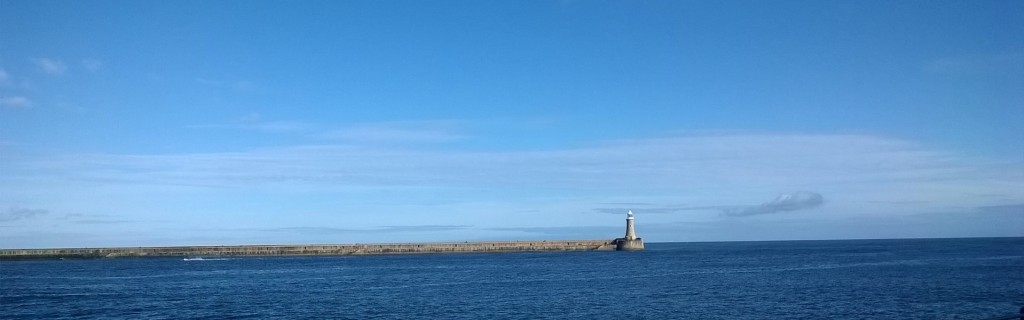 Vista looking out to see with light blue sky, deep blue sea and a lighthouse at the end of a pier