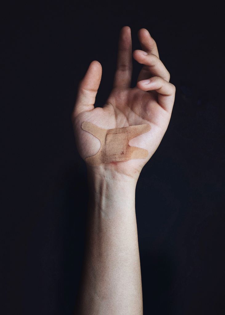 Light skinned hand, palm facing out, raised against a black background. There is an elastoplast bandage across the base of the palm.