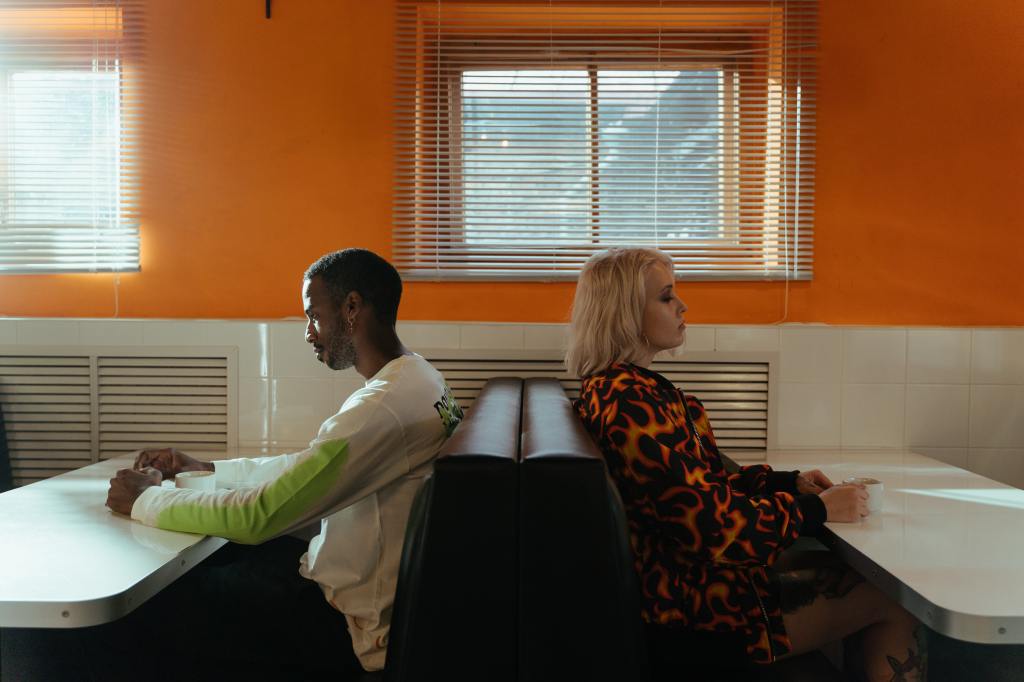 Two people sit back to back. One is a black man with short hair, the other a white woman with blonde shoulder length hair. They sit at canteen tables, with an orange wall in the background.