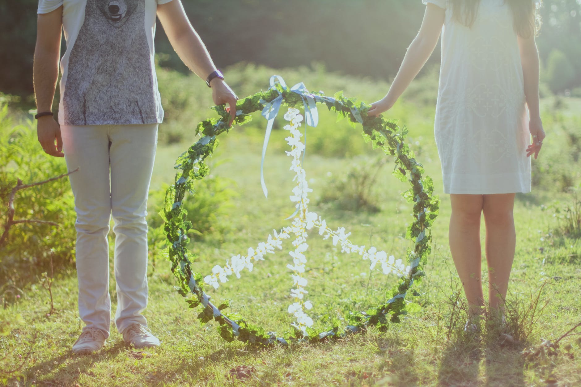 Two people (from the shoulders down) holding a hoop containing the international symbol for peace made with white flowers, surrounded by green flowers. They stand in a green field in sunlight.